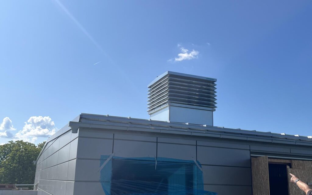 Auf dem Bild sieht man die Lamellenhaube auf dem Dach einer Schule vor strahlend blauem Himmel.