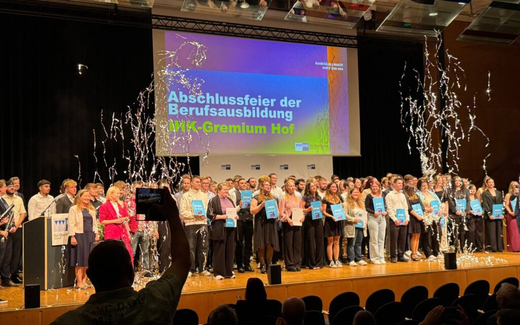 Auf dem Bild sieht man eine Bühne, auf der die Menschen stehen, die ihre Ausbildung bestanden haben und nun von der IHK HOF ihre Urkunden verliehen bekommen.
